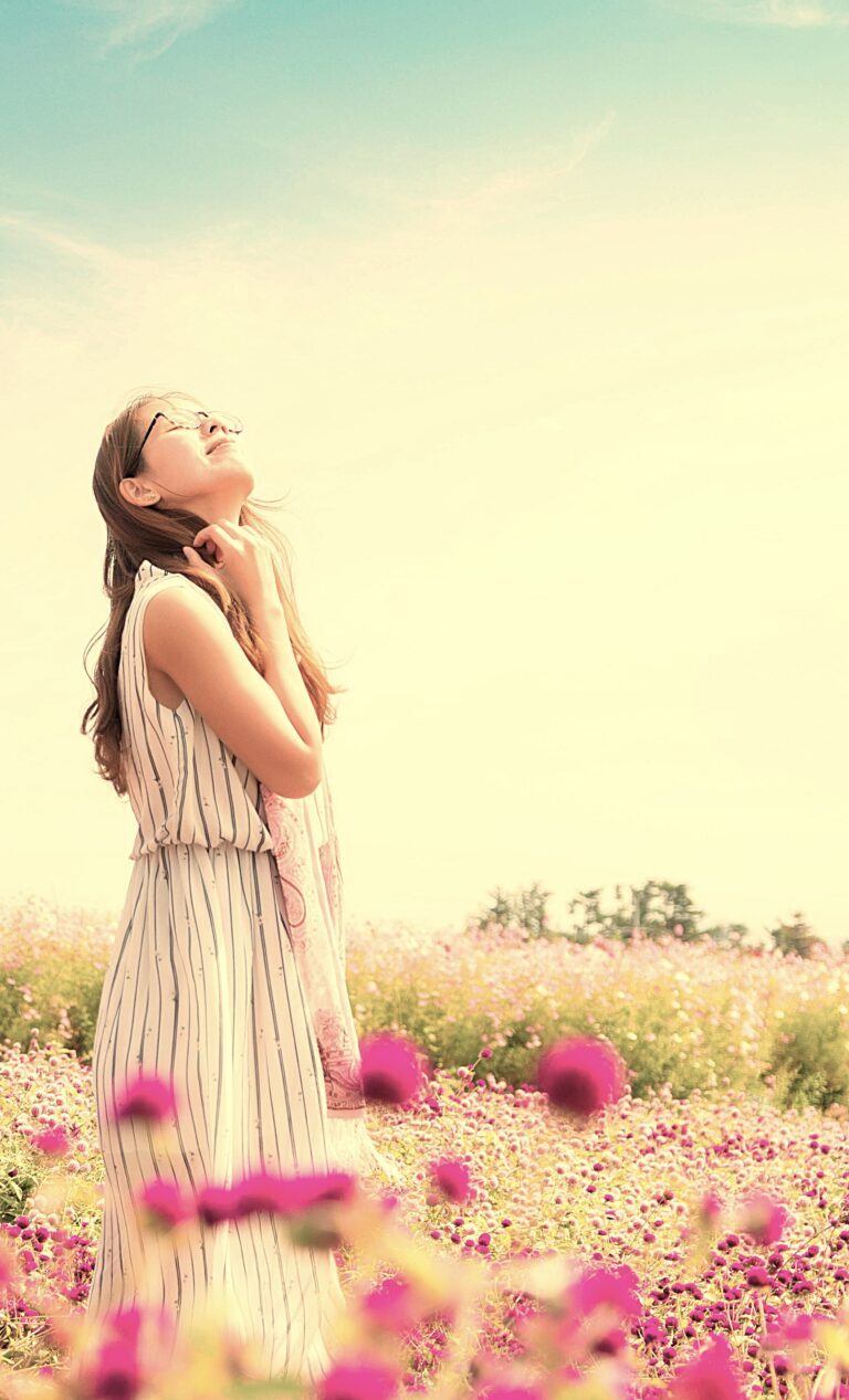 A woman standing in a field of pink and white flowers, smiling peacefully with her eyes closed, enjoying the warmth and beauty of a sunny day, symbolising mental relaxation and well-being.