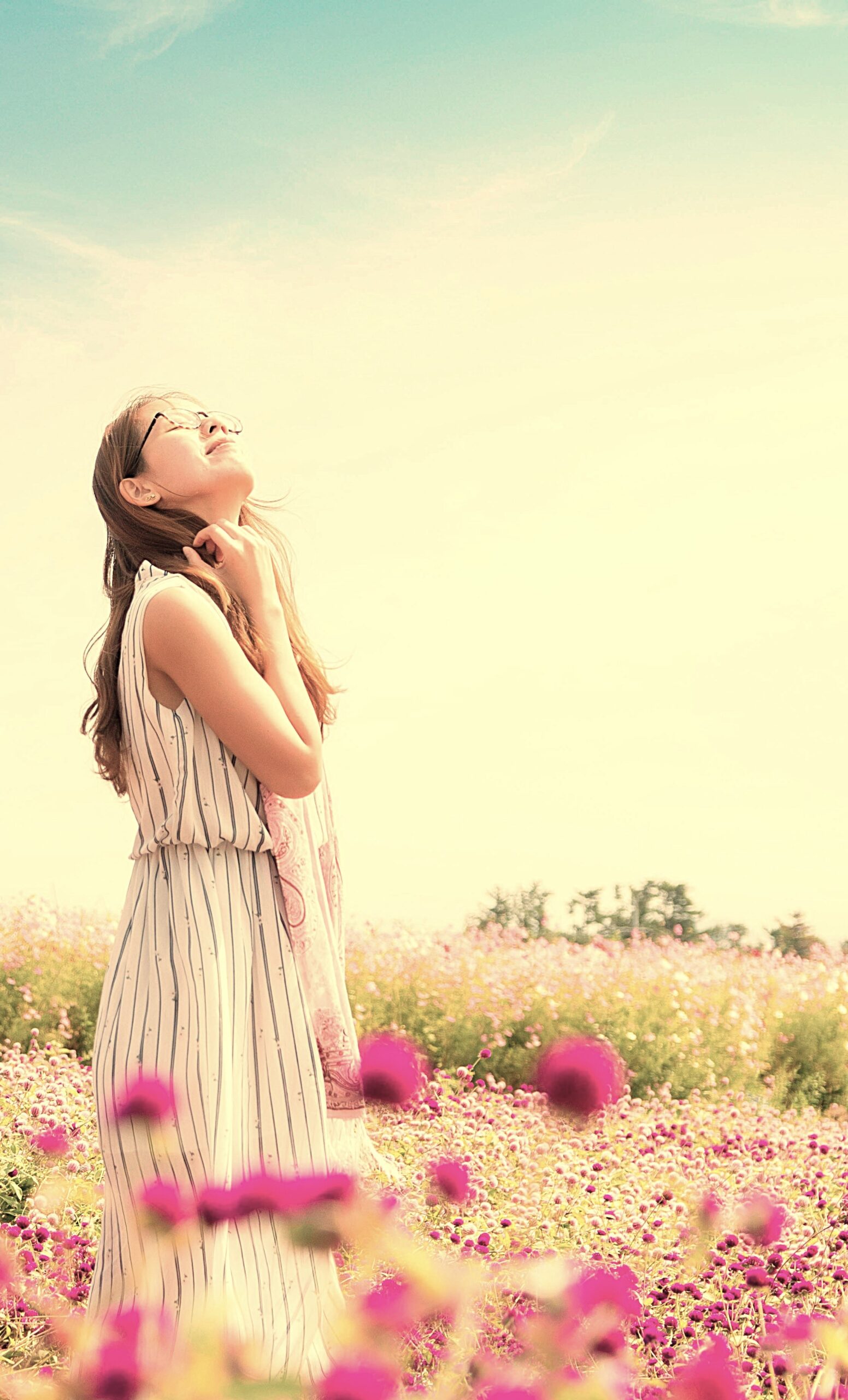 A woman standing in a field of pink and white flowers, smiling peacefully with her eyes closed, enjoying the warmth and beauty of a sunny day, symbolising mental relaxation and well-being.