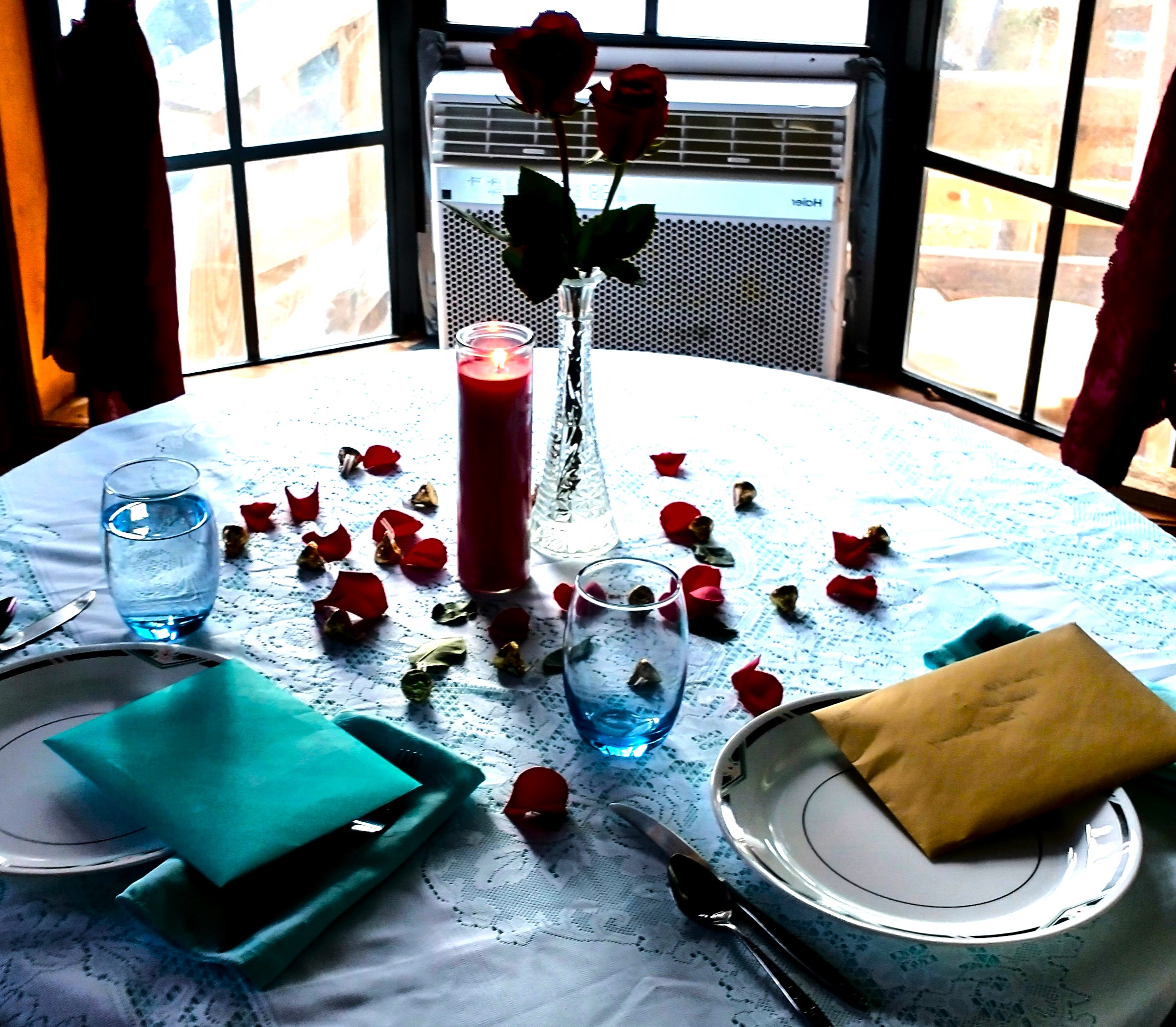 Vase with roses, candle, rose petals, and scattered chocolates on a festive dining table set for a romantic occasion. The setting suggests themes of love, intimacy, and special celebration.