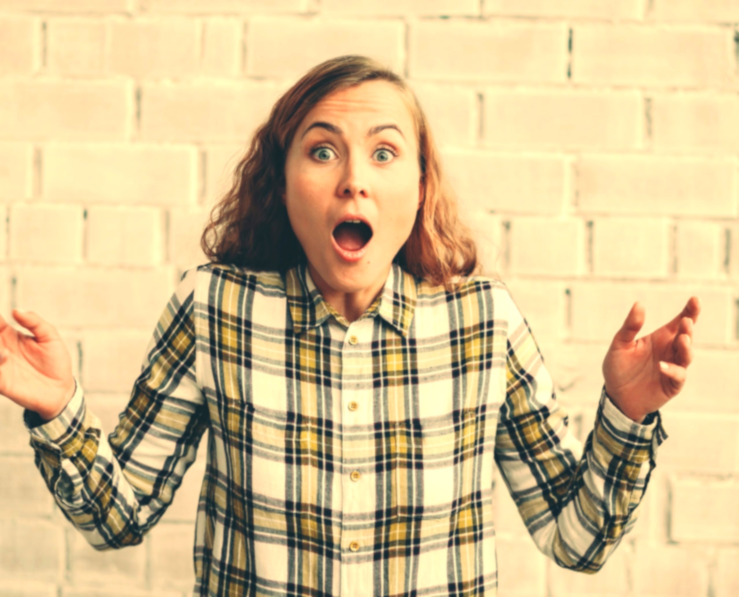 Overwhelmed woman expressing shock or frustration, standing against a light brick wall.