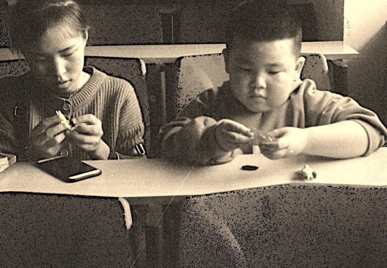 Teens sitting at a table, engaging with small objects, in a classroom setting, indicating focus and concentration.