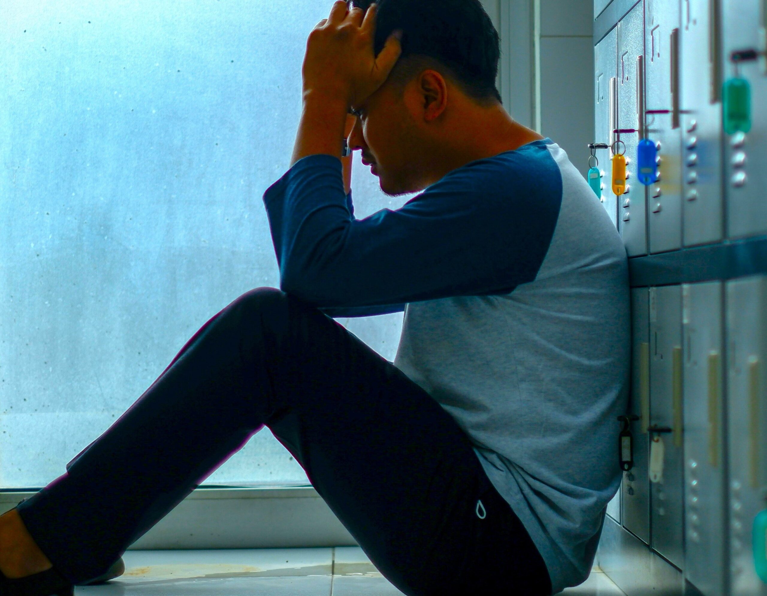 Deeply distressed young man sitting on the floor of a school corridor, holding his head in his hands, representing mental health struggles and emotional distress among teenagers.