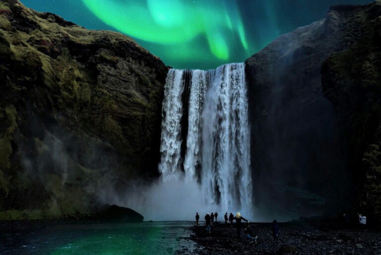 Aurora borealis above a waterfall with reflections on the water and visitors observing at the base during nighttime.