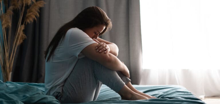 Feeling overwhelmed or struggling with emotional distress, a young woman sits alone on her bed in a moment of introspection and vulnerability.