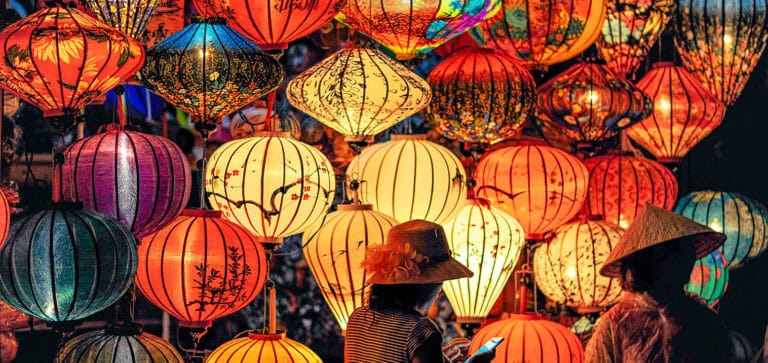 Vibrant paper lanterns at a night festival with two women wearing traditional conical hats, one using a smartphone amidst colourful, illuminated decorations creating a festive and cultural atmosphere.