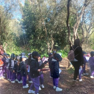 Friendliness and socialisation, children and teacher outdoors in a forest, outdoor learning, group activity, nature exploration, school trip, environmental education, early childhood development, collective experience, mental well-being, connection with nature.