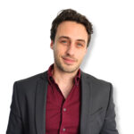 - A professional man in a dark blazer and burgundy shirt posing confidently against a plain white background.