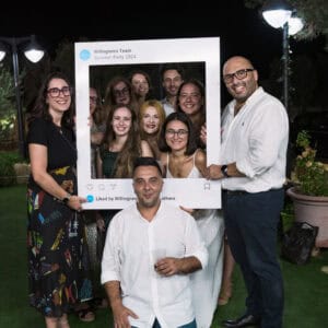 A diverse group of smiling adults at a social gathering outdoors, holding a photo frame with “Willingness Team Summer Party 2024,” reflecting camaraderie and positive mental health through social connection.
