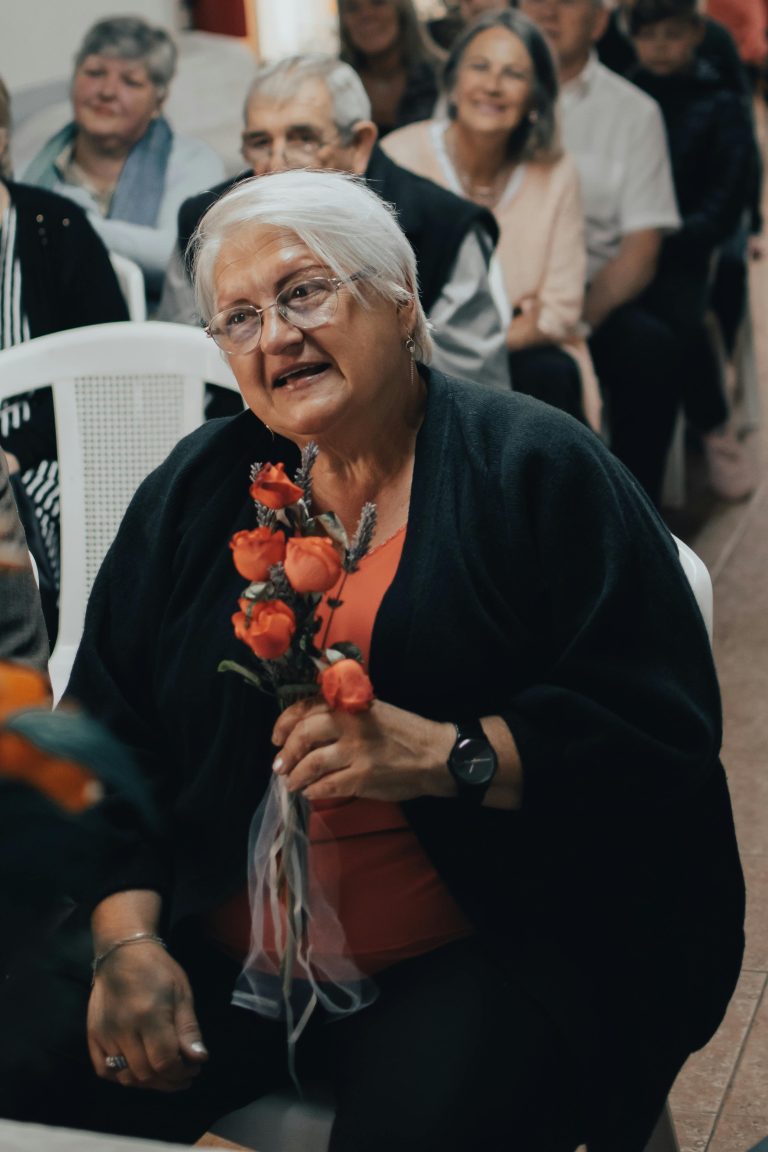 An elderly woman is shown at a family gathering, smiling happily after someone gave her roses to brighten up her day. Social relationships in old age benefit older adults immensely.