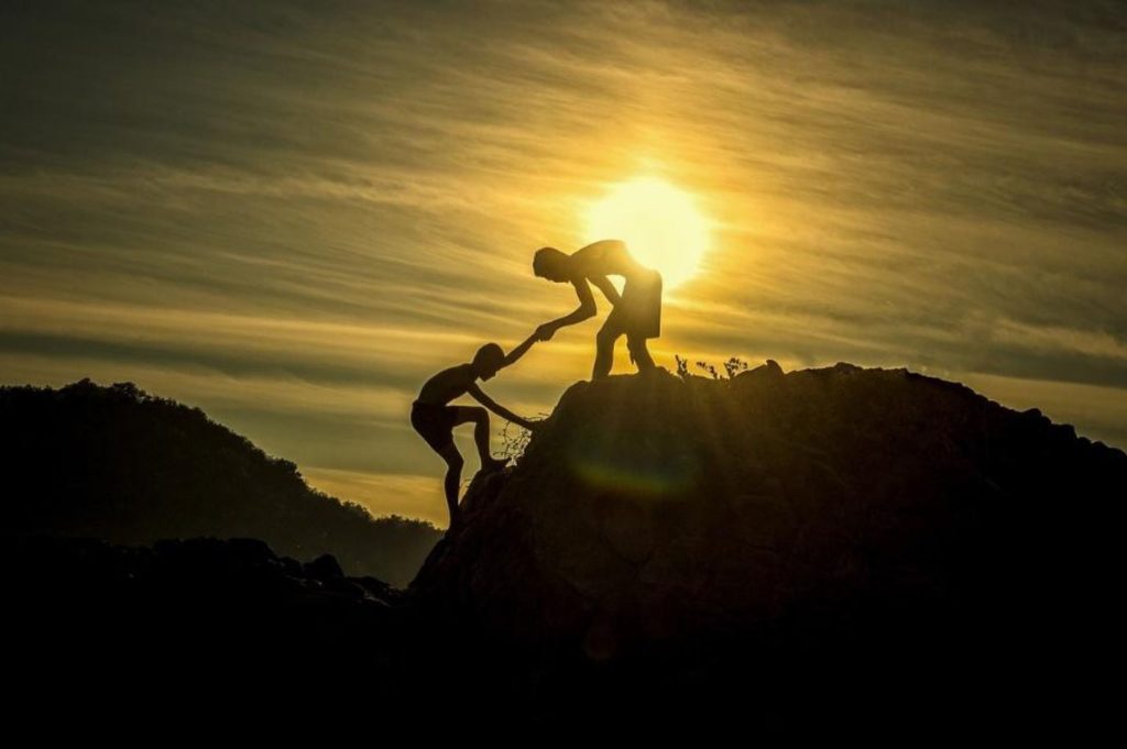 Two individuals are climbing up a mountain for the first time ever. The person at the summit is helping his friend reach the top via reaching for his friends hands, recognizing the need for help.
