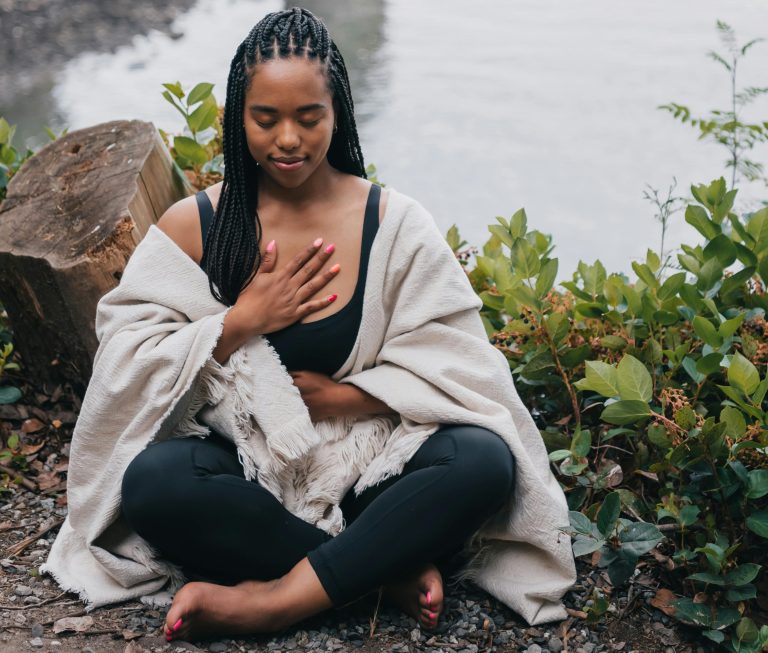 A young woman can be seen doing mindfulness at home, with some green vegetation surrounding her. This is a way of reducing the effects that trauma will have on her brain and body.
