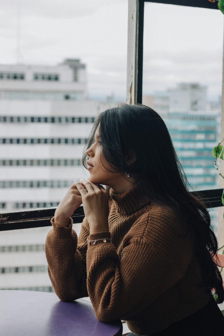 A woman in a brown sweater looking out of a window, appearing thoughtful, representing signs of trauma.
