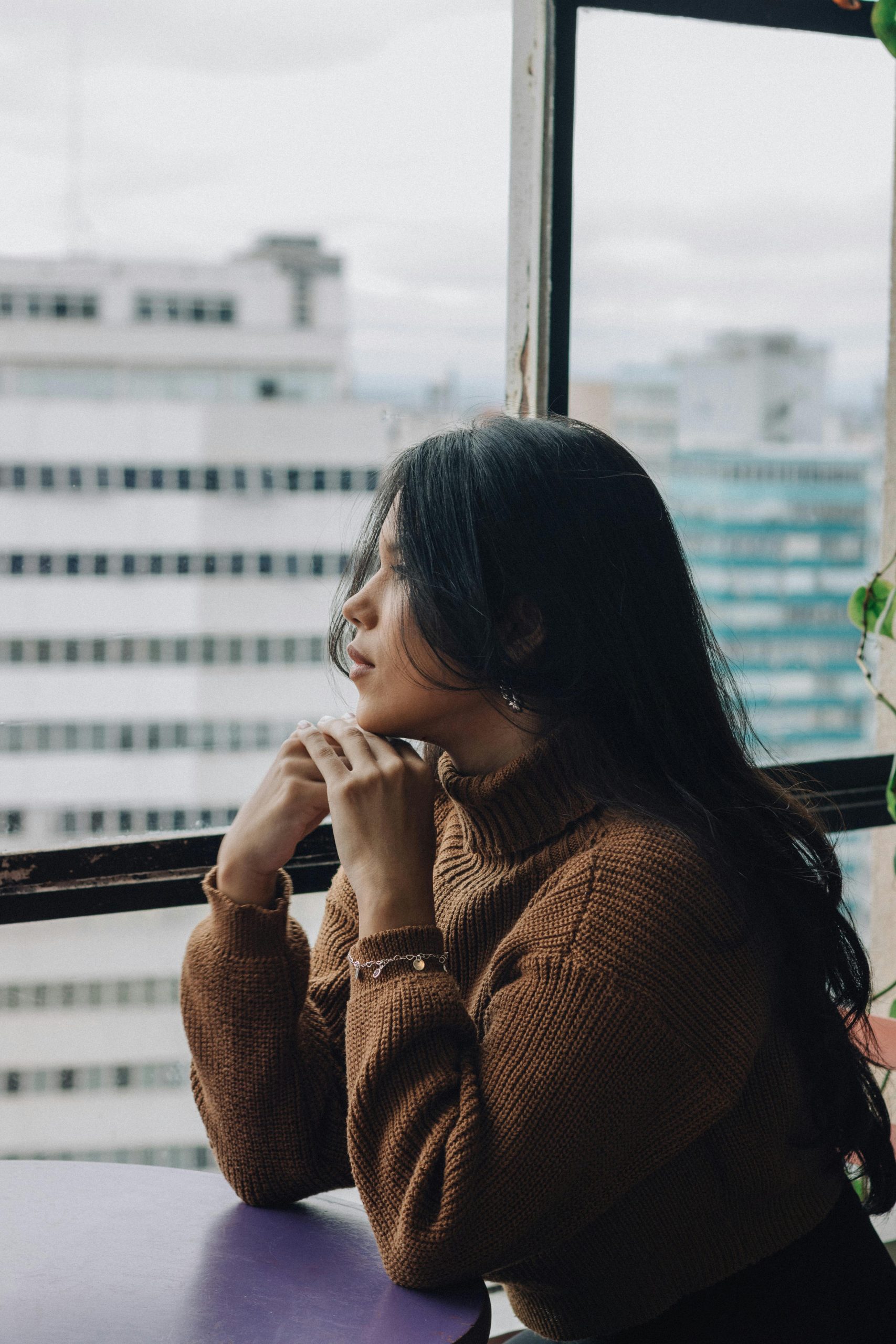 A woman in a brown sweater looking out of a window, appearing thoughtful, representing signs of trauma.