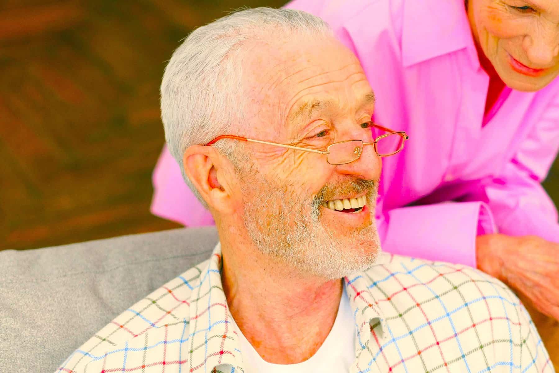 Smiling elderly man with gray hair and glasses enjoying a moment of happiness with a caregiver in a supportive setting.