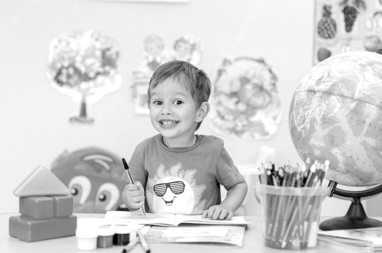 Bright young boy smiling at a desk with art supplies, a globe, and educational posters, fostering a positive learning environment that supports children's cognitive and emotional development.