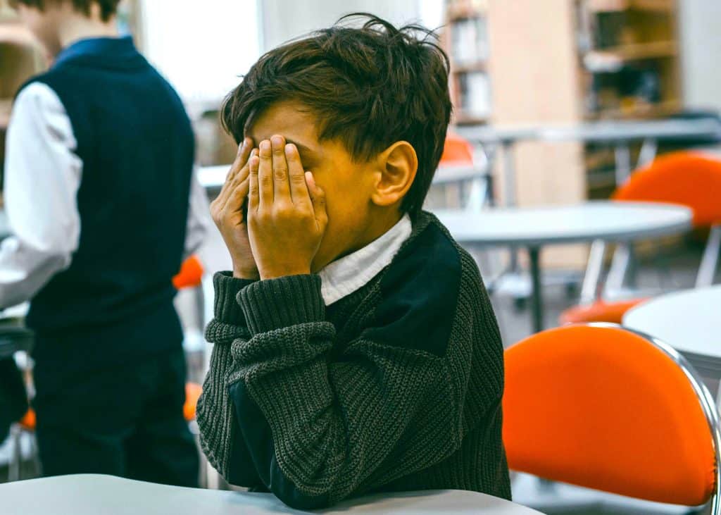 Young person with short dark hair covering their face with hands, appearing distressed in a school or library setting.