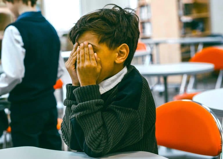 Young person with short dark hair covering their face with hands, appearing distressed in a school or library setting.