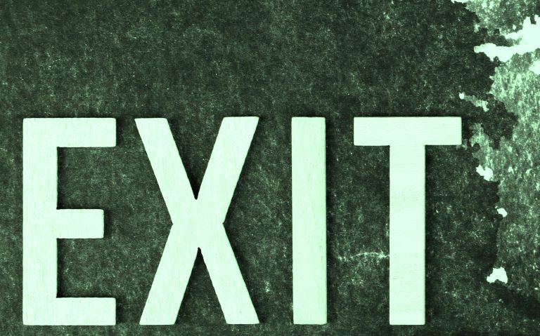 A photograph of the word "EXIT" spelled out in large, light-colored letters on a textured dark background, symbolising the concept of leaving or escaping, often used in safety signage.