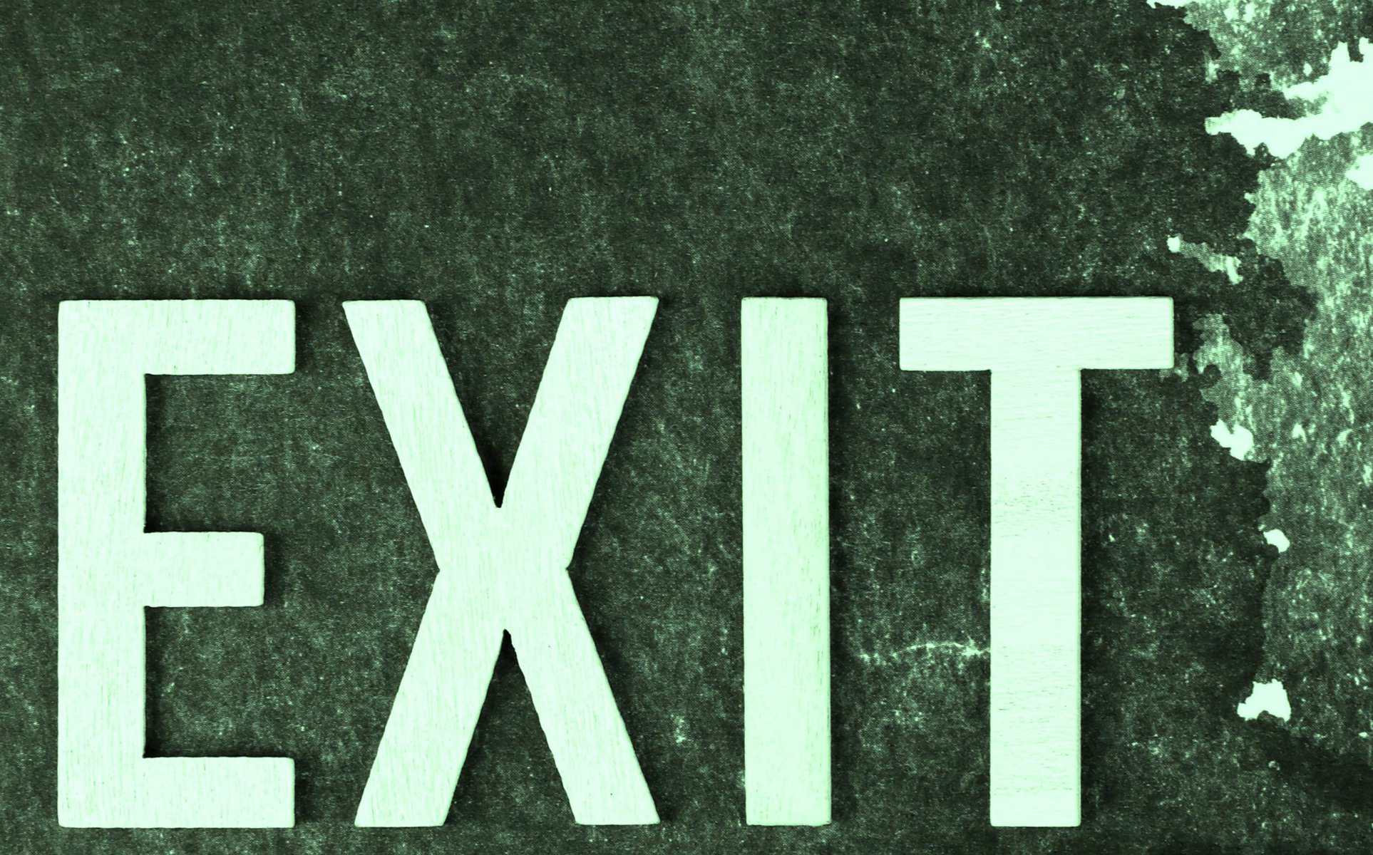 A photograph of the word "EXIT" spelled out in large, light-colored letters on a textured dark background, symbolising the concept of leaving or escaping, often used in safety signage.