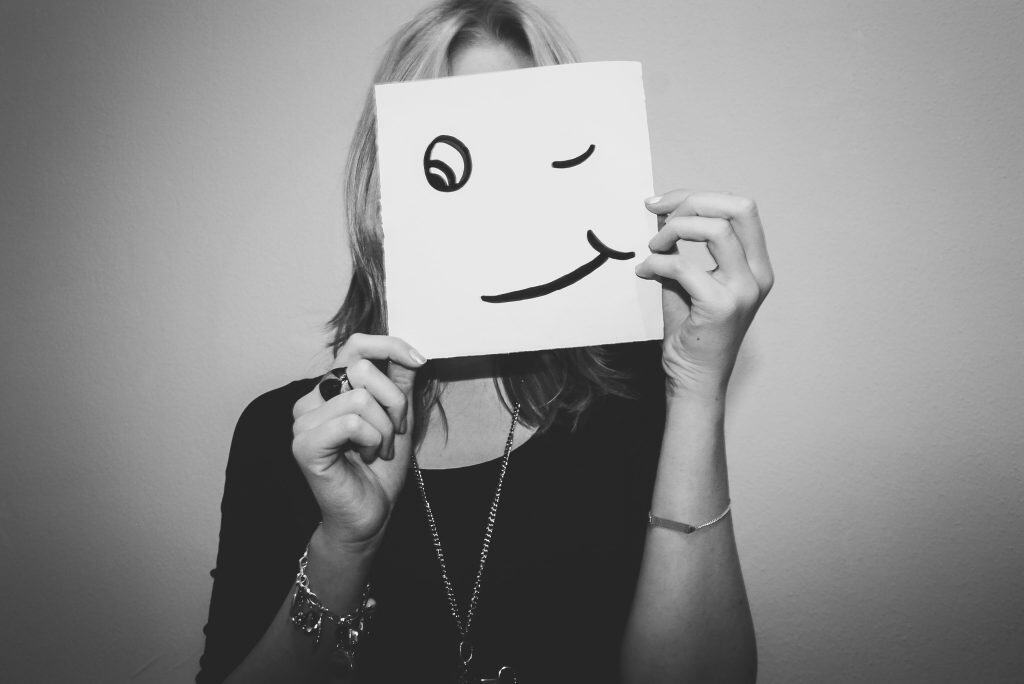 Smiling woman holding a paper with a winking face drawing in front of her face, symbolising positive mental health and emotional wellbeing.