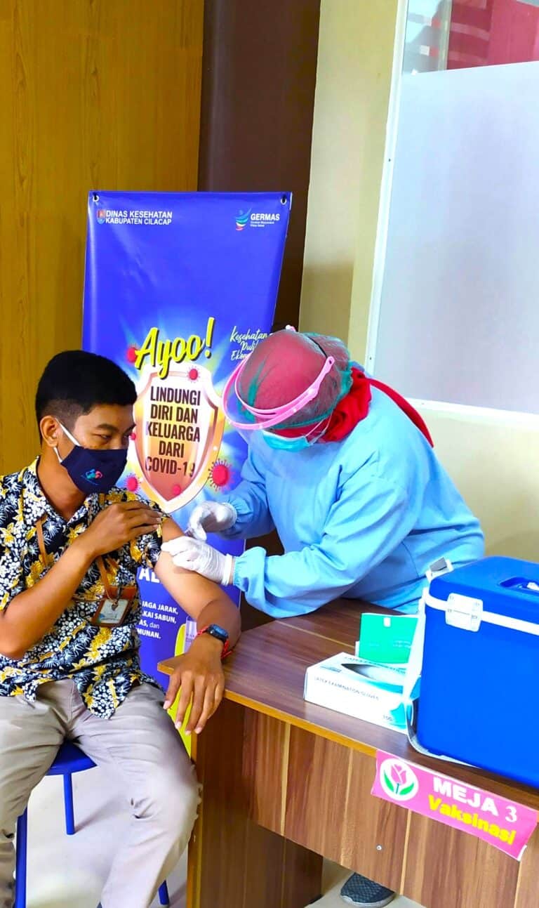 A person receiving a COVID-19 vaccine injection from a healthcare professional in a clinical setting. Both individuals are wearing masks, with the healthcare worker also wearing protective gear, demonstrating a safe vaccination process.