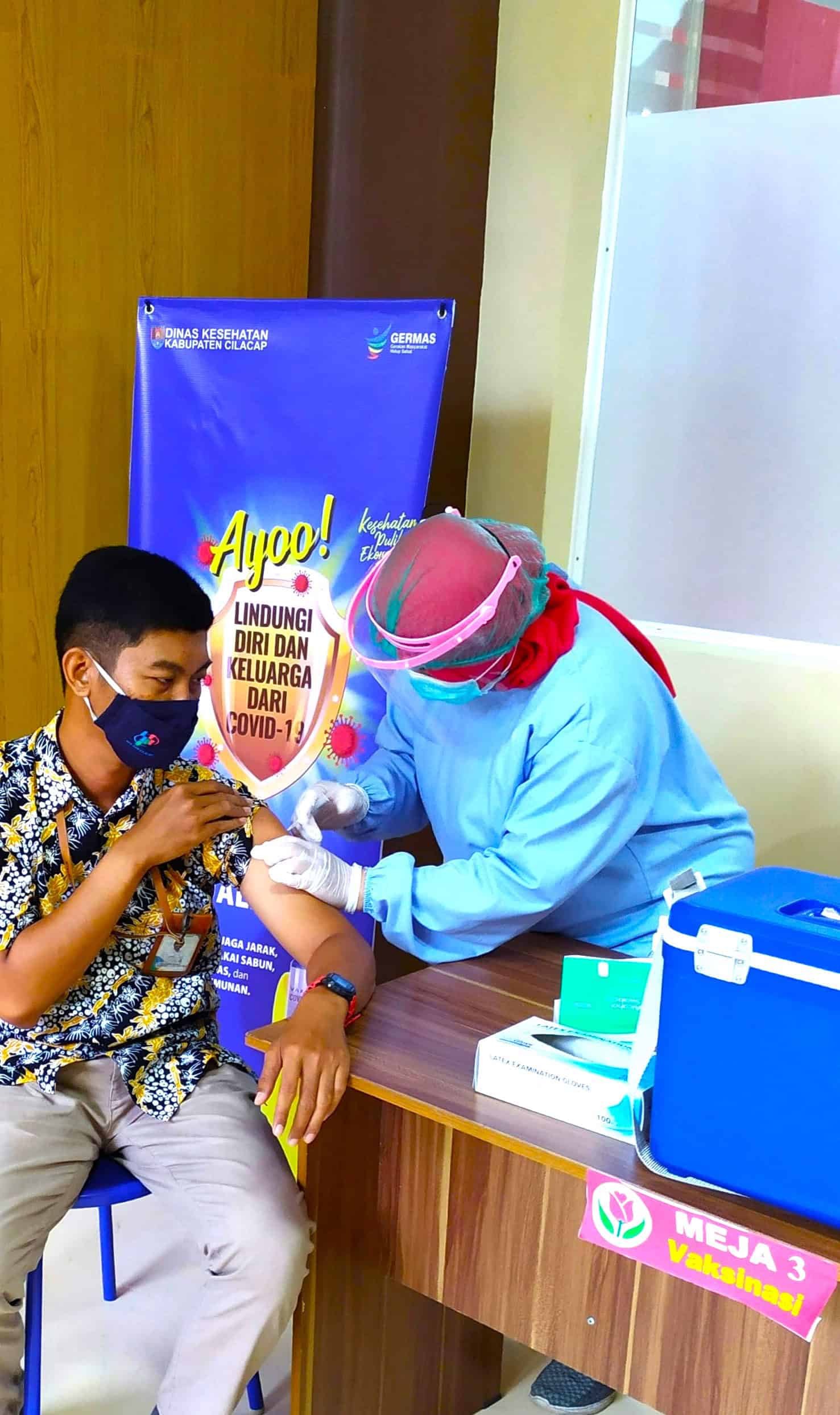 A person receiving a COVID-19 vaccine injection from a healthcare professional in a clinical setting. Both individuals are wearing masks, with the healthcare worker also wearing protective gear, demonstrating a safe vaccination process.
