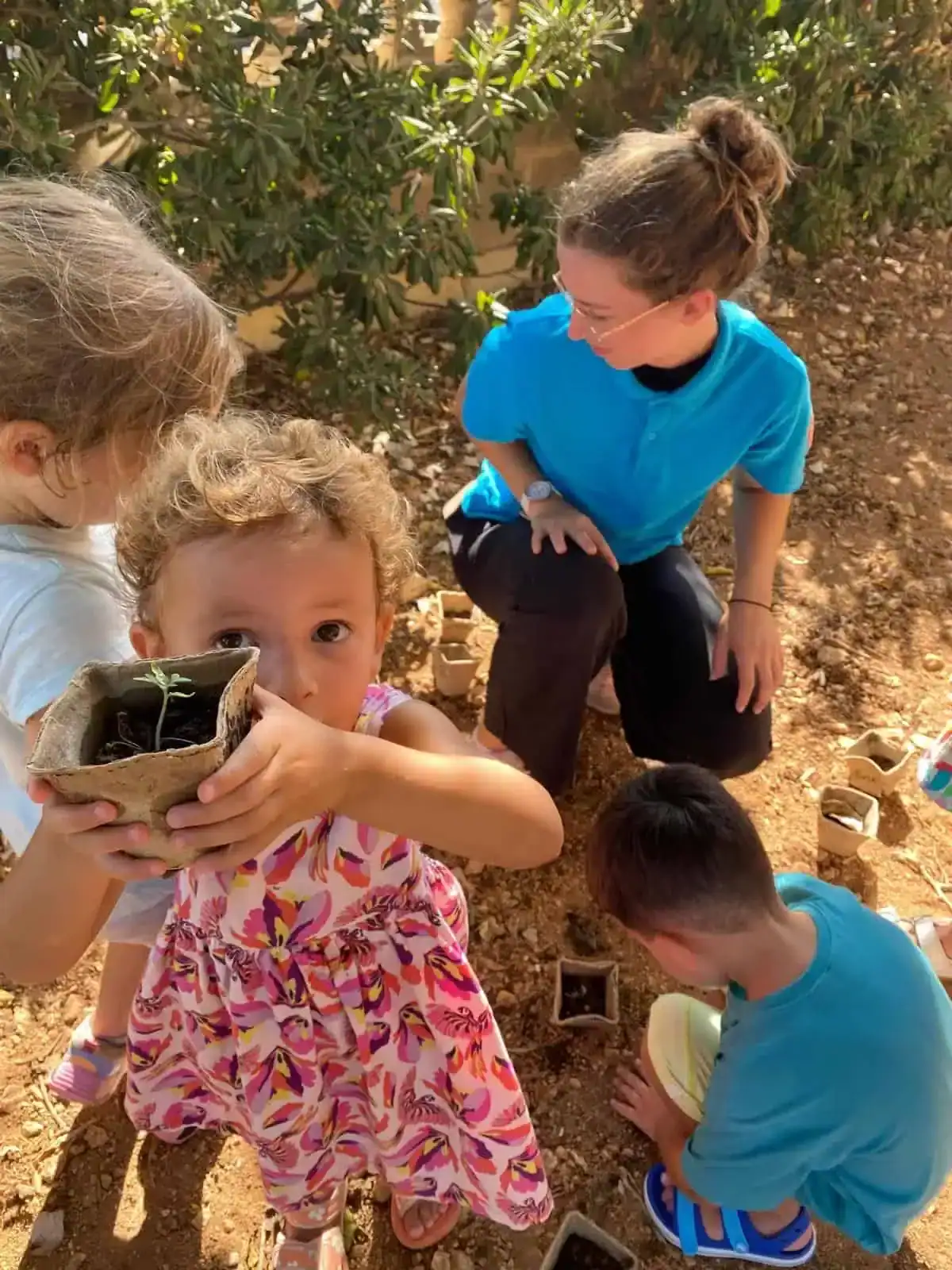 Children participating in outdoor gardening activity, promoting mental well-being and connection with nature. A caring adult supervises and engages with the children, fostering a sense of community and mindfulness.