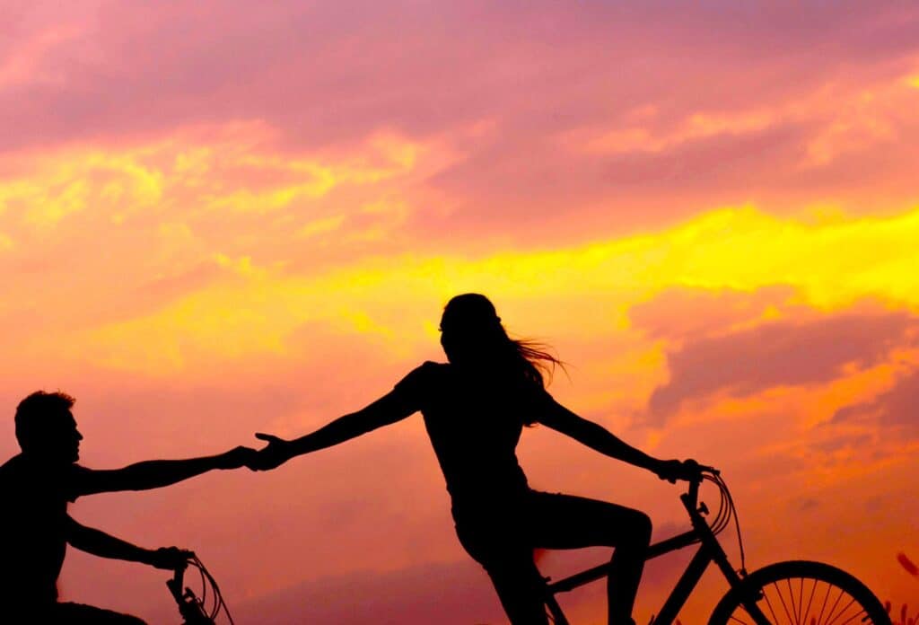 Sunset view with two people riding bikes, one helping the other by holding hands, symbolising support, connection, and emotional well-being in mental health.