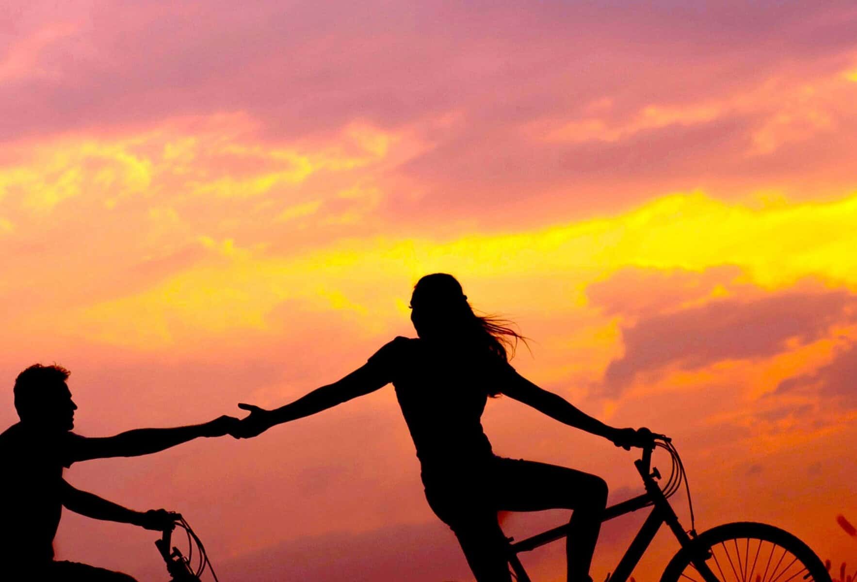 Sunset view with two people riding bikes, one helping the other by holding hands, symbolising support, connection, and emotional well-being in mental health.