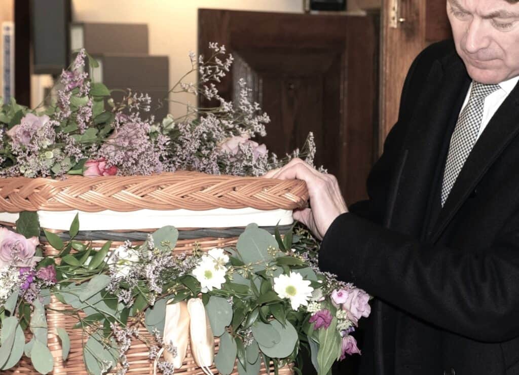 A man at a floral-covered wicker coffin, showing how Real Talk About Death helps families face grief together.