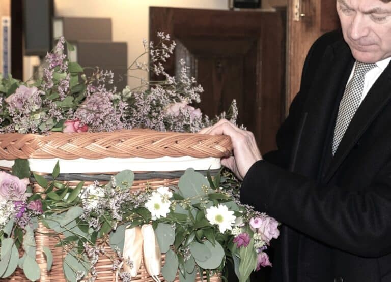 A man at a floral-covered wicker coffin, showing how Real Talk About Death helps families face grief together.