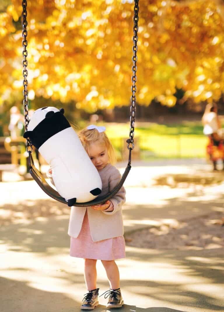 A young girl hugs a stuffed panda on a swing, showing sadness in a park during holidays from Missing Parent Feelings.
