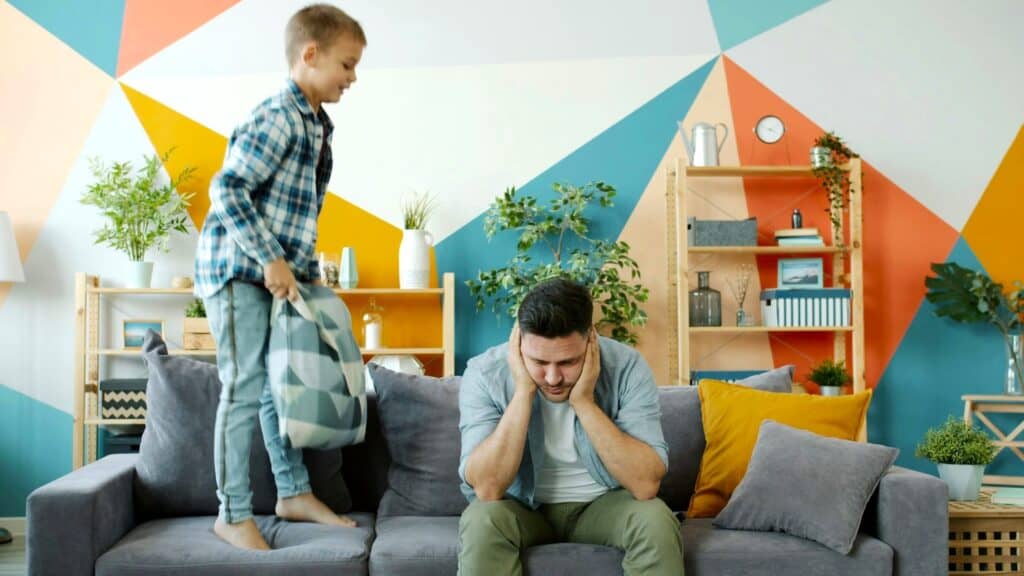 A tired father sits stressed while his child plays loudly, showing the Impact Of Parenting Stress.