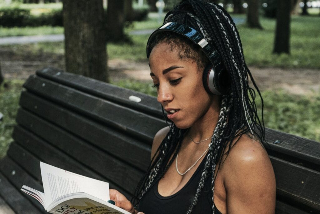 A young woman with braided hair and headphones, sitting on a park bench, engaged in reading a book, promoting relaxation and mental health awareness.