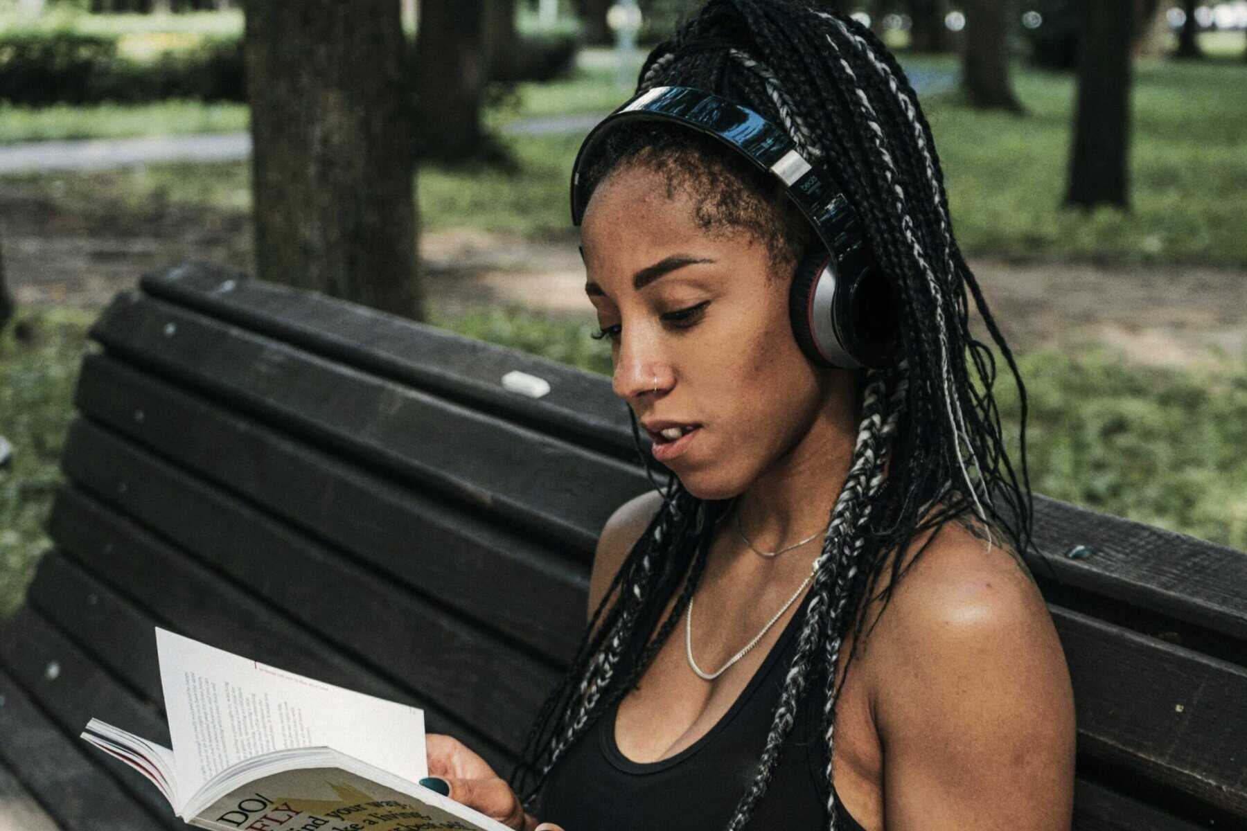 A young woman with braided hair and headphones, sitting on a park bench, engaged in reading a book, promoting relaxation and mental health awareness.