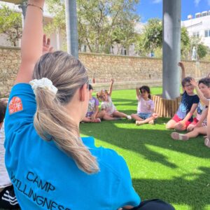 Children participating in a summer camp activity outdoors, sitting on the grass and engaging in a group discussion led by an instructor.