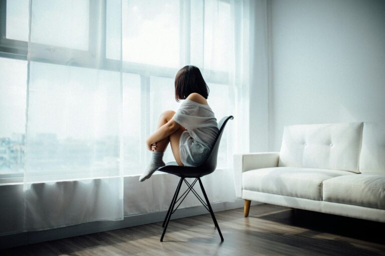 A young girl sits alone on a chair by a large window, gazing outside, symbolising introspection and emotional reflection.
