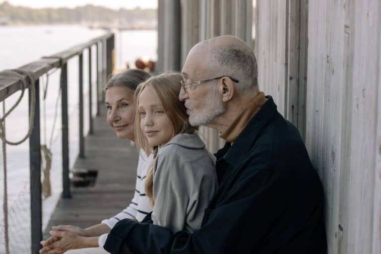 An elderly man with glasses and two young girls sitting on a wooden dock by the water, sharing a peaceful moment together.